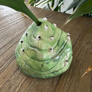 May include: A green ceramic vase with a unique, organic shape and white dots. The vase is designed to look like a rock or a mountain.