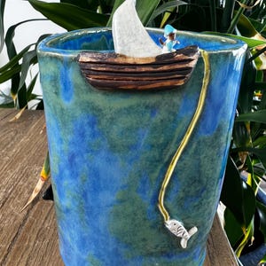 May include: A ceramic planter with a blue and green glaze. A small boat with a white sail and a tiny figure is attached to the rim. A gold-coloured line extends from the boat to a silver fish.