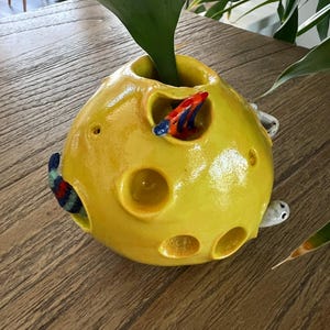 May include: A yellow ceramic vase shaped like a moon with holes and a green plant growing out of the top. There are small ceramic fish and snails on the vase.