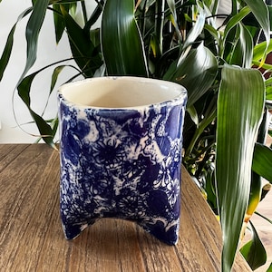 May include: A blue and white ceramic planter with a floral design and three legs. The planter is sitting on a wooden surface.