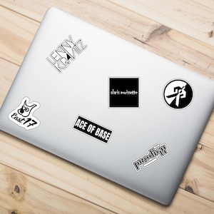 May include: A white laptop covered in various black and white stickers. Stickers include band names such as "Lenny Kravitz", "Ace of Base", and "Prodigy". The laptop is on a light brown wooden surface.