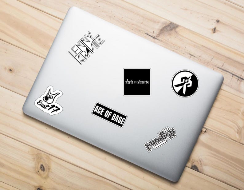 May include: A white laptop covered in various black and white stickers. Stickers include band names such as "Lenny Kravitz", "Ace of Base", and "Prodigy". The laptop is on a light brown wooden surface.