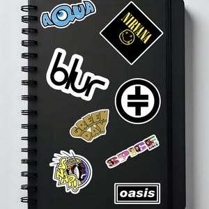 May include: Black notebook with spiral binding, featuring stickers of the bands Aqua, Blur, Green Day, Nirvana, Spice Girls, Snap!, Oasis, and a black and white symbol. The stickers are in various colors and designs.
