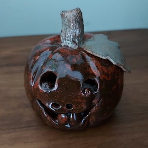May include: A brown ceramic pumpkin with a carved face and a green stem. The pumpkin has a glossy finish and is decorated with red and brown speckles.
