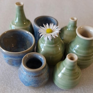 May include: A selection of small, handmade ceramic vases and bowls in shades of blue and green. A white daisy sits in one of the vases. The pottery has a glossy finish and various shapes, including a tall, slender vase and a small, round bud vase.