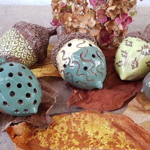 May include: Five ceramic acorn shaped ornaments with different designs. The acorns are brown, green, and yellow with brown, green, and white patterns. The acorns are sitting on a bed of brown and yellow leaves.