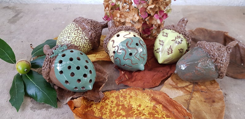 May include: Five ceramic acorn shaped ornaments with different designs. The acorns are brown, green, and yellow with brown, green, and white patterns. The acorns are sitting on a bed of brown and yellow leaves.