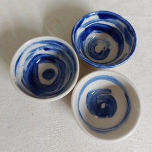May include: Three small white ceramic bowls with blue swirl designs. The bowls are arranged in a triangular pattern on a white surface.
