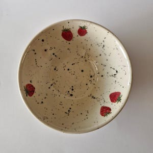 May include: A round ceramic bowl with a cream-coloured base featuring a speckled black pattern throughout the interior. The bowl is decorated with small, hand-painted red strawberries with green leaves scattered across the inner surface.