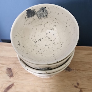 May include: A stack of three off-white ceramic bowls featuring a speckled grey pattern and a black line-art illustration of two sheep near the inner rim. The bowls have a smooth, rounded shape and a consistent design across the entire set.