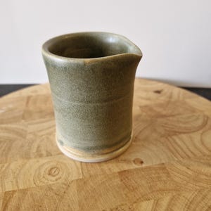 May include: A small, green ceramic jug with a white rim. The jug is sitting on a wooden surface.