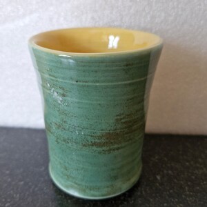 May include: A handmade ceramic tumbler, green on the outside and yellow inside. The tumbler is cylindrical with a flared rim. The green glaze has a textured, rustic look, with brown speckling. The interior is smooth, glossy yellow, giving a contrast of colours.