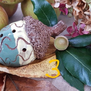 May include: A ceramic acorn whistle painted in brown, green and white. The whistle has three finger holes and is decorated with a swirling pattern. The acorn is sitting on a brown leaf with the number 3 written on it.