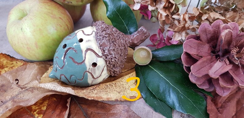 May include: A ceramic acorn whistle painted in brown, green and white. The whistle has three finger holes and is decorated with a swirling pattern. The acorn is sitting on a brown leaf with the number 3 written on it.