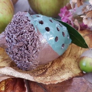 May include: A ceramic acorn with a brown and green glaze. The acorn has a textured surface and a hole in the top. The acorn is sitting on a bed of brown leaves.