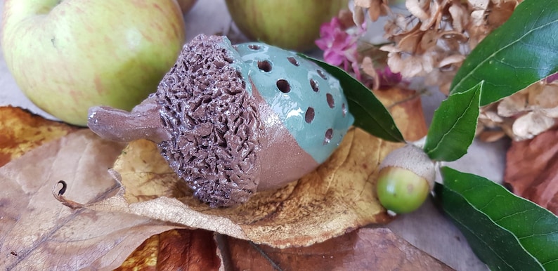 May include: A ceramic acorn with a brown and green glaze. The acorn has a textured surface and a hole in the top. The acorn is sitting on a bed of brown leaves.