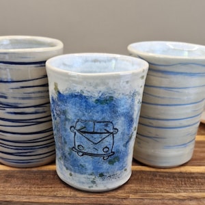 May include: Three handmade ceramic tumblers in shades of blue and white. One has a blue abstract design, another has blue and white stripes, and the third features a blue painted background with a black van illustration. Each tumbler is unique.
