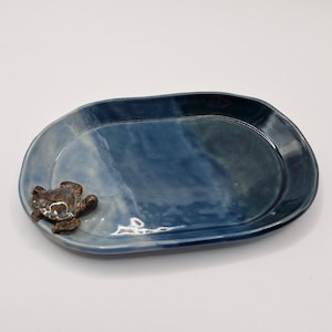 May include: An oval-shaped ceramic tray with a glossy, variegated blue glaze. The surface features a smooth, slightly uneven rim. A small, three-dimensional brown and tan turtle figure is attached to the lower left edge of the tray.
