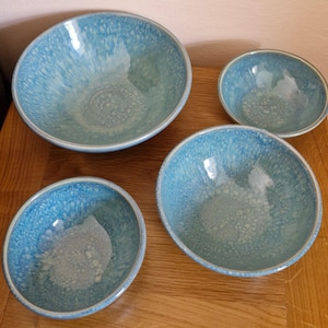 May include: A set of four ceramic bowls in different sizes. They feature a light blue glaze with a speckled design. The rims are a darker shade of blue. The bowls are displayed on a wooden surface.
