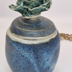 May include: A blue and green ceramic jar with a lid shaped like a flower. The flower is green with gold accents.