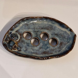 May include: A handmade ceramic soap dish with a blue and brown glaze. The oval dish features four circular drainage holes and a textured surface. The edges are a darker brown, and the design is organic and unique, suitable for the bathroom.