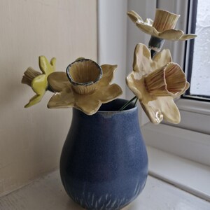 May include: A navy blue ceramic vase displays ceramic daffodil sculptures. The daffodils have yellow petals and brown centers. The vase is positioned on a white surface near a window.