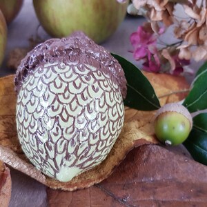 May include: A hand-painted ceramic acorn with a brown cap and a green body with brown swirl designs. The acorn is sitting on a brown leaf with a green leaf and a small acorn in the background.