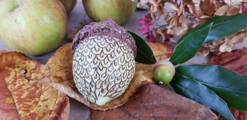May include: A hand-painted ceramic acorn with a brown cap and a green body with brown swirl designs. The acorn is sitting on a brown leaf with a green leaf and a small acorn in the background.