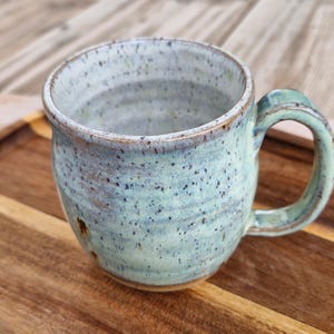 May include: A ceramic mug with a speckled light blue and green glaze. The mug features a rounded body, a sturdy handle, and a textured surface with visible brown flecks throughout the finish. The rim has a subtle brown edge, and the base is unglazed.