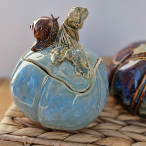 May include: A blue ceramic pumpkin-shaped container with a snail figurine and green leaf detail. The container is on a woven surface. The pumpkin-shaped container is a decorative item.