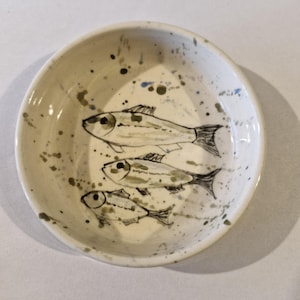 May include: A small, off-white ceramic bowl featuring a hand-painted design of three fish. The fish are sketched in black and grey, with speckled accents of green and blue. The bowl's rim is slightly curved.