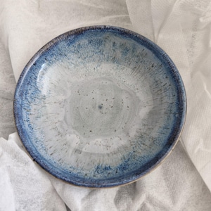 May include: A round ceramic bowl featuring a reactive glaze with a deep blue rim that transitions into a speckled light grey and white centre. The surface displays a subtle texture with fine dark flecks throughout the interior basin.