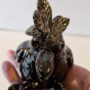 May include: A glossy, dark brown ceramic bird figurine with large, expressive eyes and a leafy crest. The bird is in a seated position, with its feet and beak clearly defined. The figurine has a smooth, reflective surface.