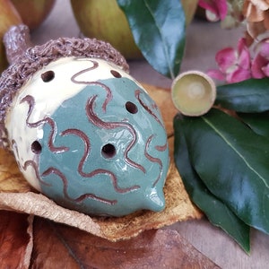 May include: A ceramic acorn ornament with a brown cap and a green and white body with brown swirl designs. The acorn has several holes in the body. The ornament is surrounded by autumn leaves, a pine cone, and bay leaves.