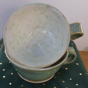 May include: Two handmade ceramic soup bowls stacked. The bowls are light green with a speckled pattern and a handle. They sit on a dark green surface. Each bowl is approximately 15 cm in diameter.