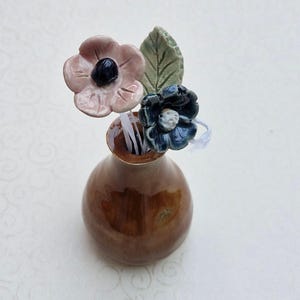 May include: A small, brown, glazed ceramic vase containing three decorative ceramic stems. One stem features a light pink flower with a dark centre, another a dark blue flower with a white centre, and the third is a textured green leaf.