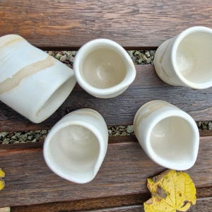 May include: Five white ceramic cups with a light brown glaze. The cups are all different shapes and sizes, and they are arranged in a row on a wooden surface.