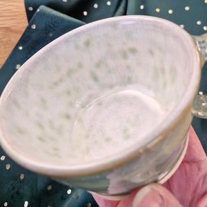 May include: A ceramic mug with a speckled, pale green and white interior. The mug has a rounded shape, a handle, and a cream-coloured rim. The mug is set against a dark green background.