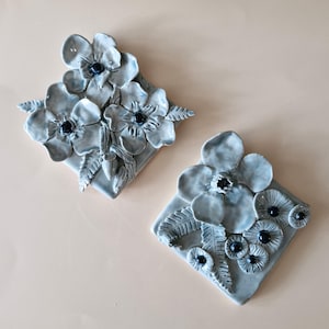 May include: Two ceramic wall art pieces with a floral design. The light blue pieces have dark blue centres and are set against a light beige backdrop. The flowers, with detailed petals and leaves, are the main focus of the artwork.