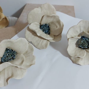 May include: Handmade ceramic flower sculptures in a creamy off-white colour with textured petals. The centres of the flowers are a contrasting blue-green hue. The flowers are arranged on a white surface, showcasing their delicate details.