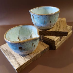 May include: Two ceramic bowls with a light blue glaze and brown speckles, each with a spout. They sit on small, square wooden coasters. The bowls have a rustic, handmade appearance.