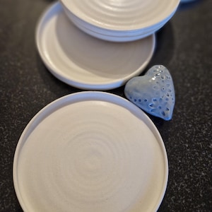 May include: A set of four white ceramic plates with a subtle swirl design. The plates are stacked on top of each other, with a blue heart-shaped decorative object in the foreground.