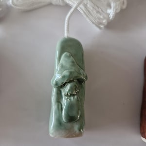 May include: A light green ceramic pull chain with a mushroom design. The mushroom has a textured cap and a small pouch. A white cord is attached for hanging. The ceramic piece is approximately 5 cm long.