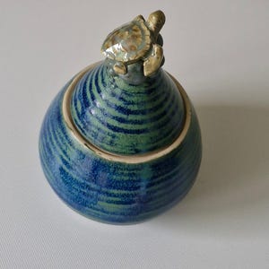 May include: A round ceramic jar with a lid featuring a small sculpted turtle on top. The jar and lid have a glossy glaze with horizontal bands of deep blue and sage green, showing a textured, ribbed surface throughout the entire exterior.