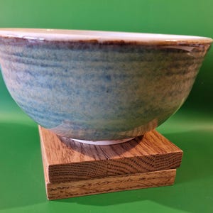 May include: A handcrafted ceramic bowl with a blue and brown glaze. The bowl sits on a small, square wooden stand. The bowl's interior is white, and the exterior has a textured, mottled appearance.