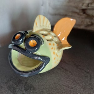 May include: A ceramic fish figurine with a green and yellow glaze. The fish has large, black eyes with orange pupils and a wide, open mouth. The fish has a small, orange fin on its back.