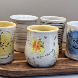 May include: Five handcrafted ceramic mugs with varied designs. The mugs display floral and abstract patterns in yellow, blue, and white, presented on a wooden tray. Each mug has a unique design, suitable for beverages.