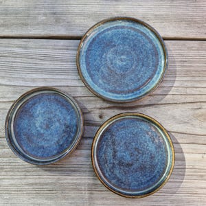 May include: Three circular ceramic plates featuring a mottled blue and brown glaze with a smooth, glossy finish. The plates vary slightly in size, each displaying a distinct reactive glaze pattern that blends deep navy tones with lighter blue hues.