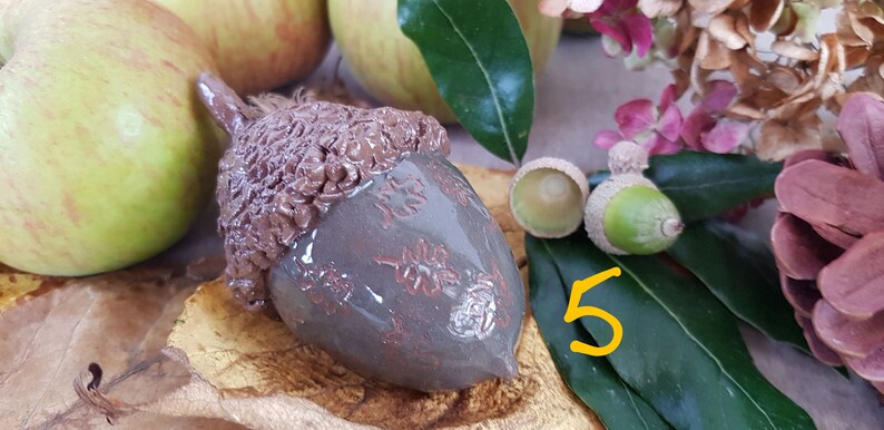 May include: A ceramic acorn ornament with a brown and grey glaze. The acorn is decorated with a leaf pattern and has a textured cap.