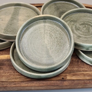 May include: Six small, round, sage green ceramic plates with a raised rim, arranged on a dark brown, oval wooden tray. The plates, with a textured surface, are presented to highlight their earthy tones and simple design. The tray is oval-shaped.